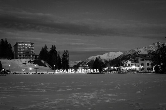 Scenic view of frozen and snow covered Etang Grenon (Lac Grenon) with illuminated lettering "Crans Montana", Switzerland against mountains and blue sky with clouds in black and white