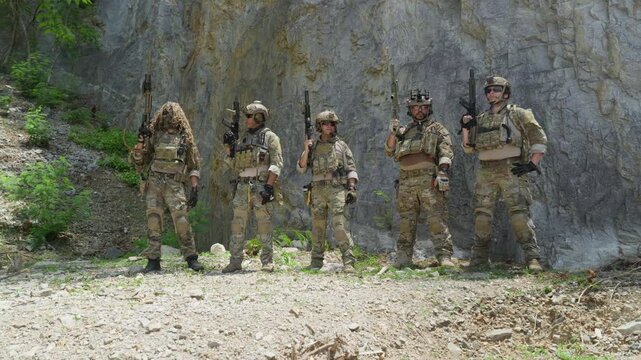 Squad of five soldiers in camouflage uniform standing in lineup formation during military tactical operation outdoor, special forces operators holding rifle weapons near rock cliff