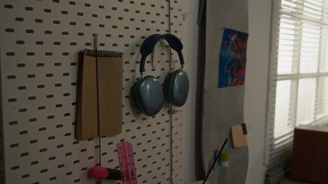 Blue wireless headphones and notebook hanging on pegboard organizer in student room. Footage represents modern study space, ideal for educational marketing or remote work campaigns
