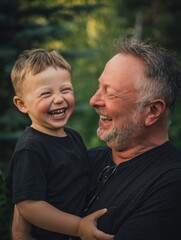 Fototapeta premium Father's Day. Older father cradling laughing baby against chest, gray in hair, black shirt, forest backdrop. Late parenthood, mature dad joy, tender natural portrait, older age fatherhood..