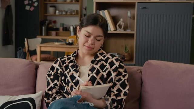 Portrait of young Hispanic female college or university student sitting on sofa, writing in notebook, then looking at camera and smiling. Youth lifestyle, budgeting, or remote learning concepts