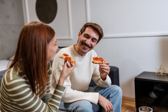 A happy young couple eats pepperoni pizza together on a sofa at home.