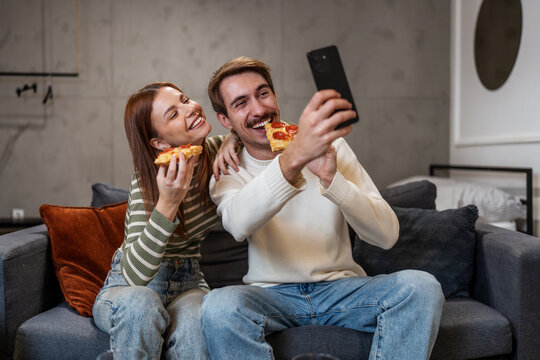 A cheerful young couple takes a selfie on a smartphone while enjoying pizza slices on a cozy living room sofa.