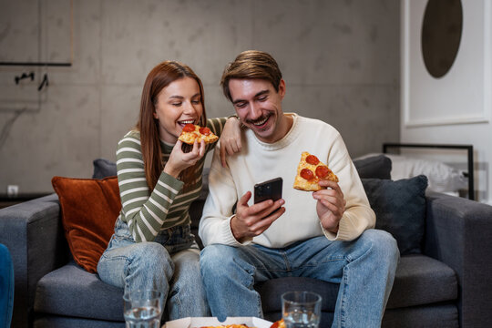 A cheerful young couple enjoys eating pepperoni pizza while looking at a smartphone together on a cozy sofa in their modern living room.