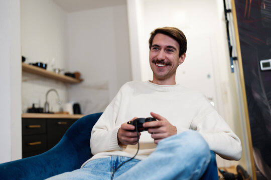 A happy young man playing video games with a controller while sitting on a comfortable armchair in his apartment.
