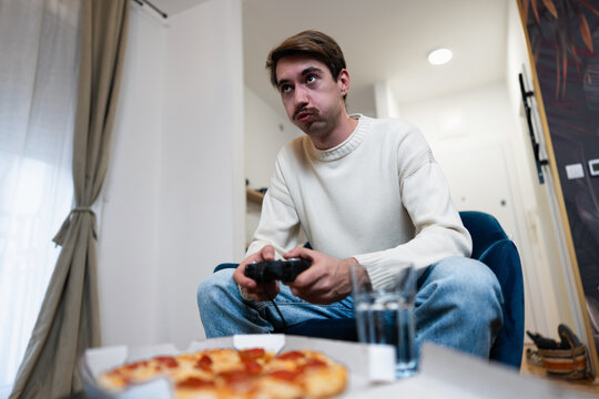Focused gamer playing video games on a console while sitting in a living room with pizza nearby.