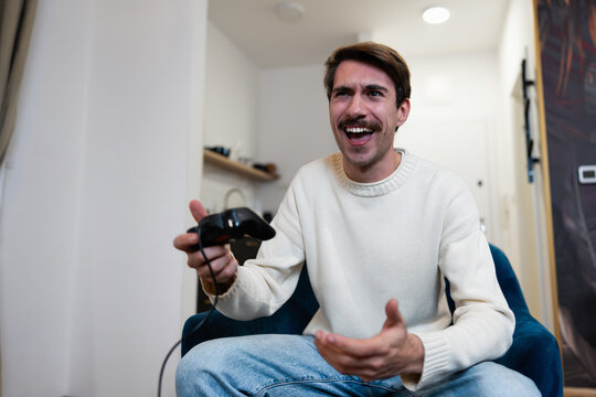A happy young man playing video games with a controller while sitting on a comfortable armchair in his apartment.