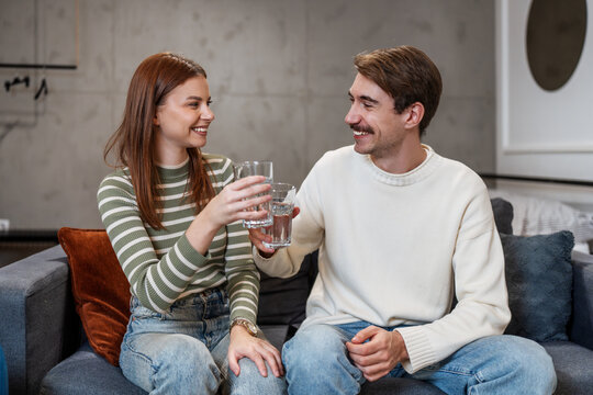 Happy couple holding glasses of water and clinking them together while sitting on a comfortable sofa in their modern living room, celebrating healthy lifestyle habits.