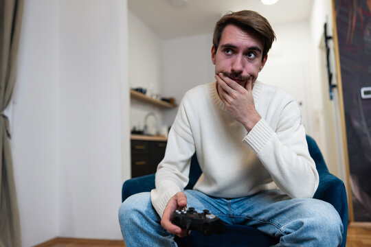 A concentrated gamer sitting in an armchair holding a controller, looking pensive and thinking about his next move.