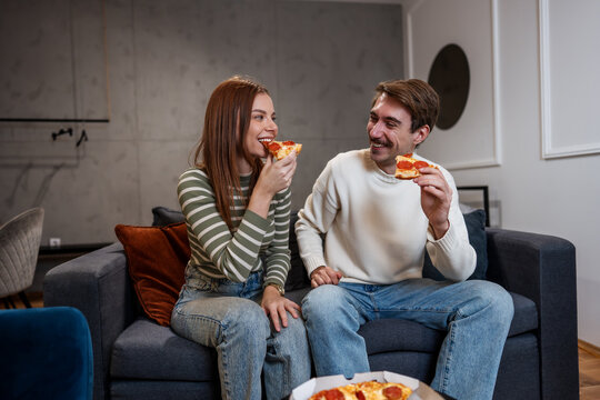 A happy young couple eats pepperoni pizza together on a sofa at home.