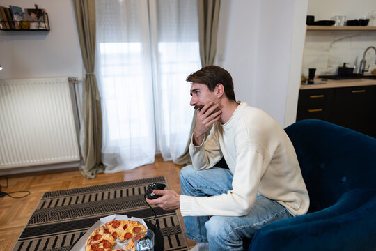 Focused gamer playing video games on a console while sitting in a living room with pizza nearby.