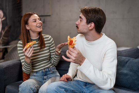 A happy young couple eats pepperoni pizza together on a sofa at home.
