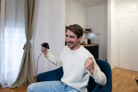 A happy young man playing video games with a controller while sitting on a comfortable armchair in his apartment.