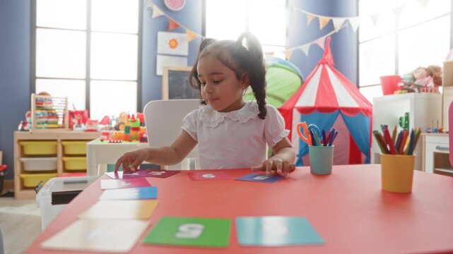 Girl placing number cards with hand on a red activity table, sitting in a bright playroom with tent and toy storage in building; concentration learning.