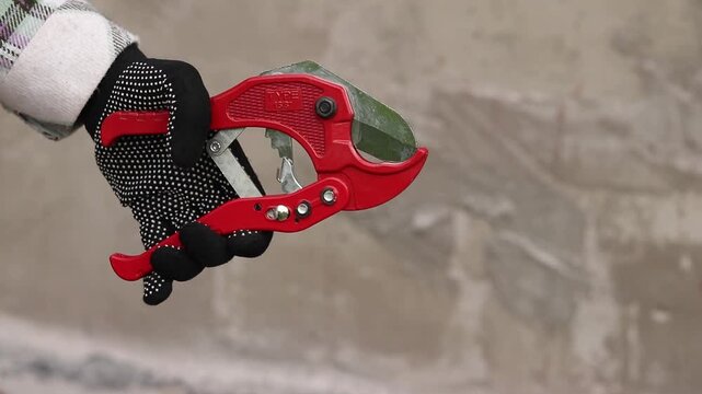 A hand in a work glove holds a red pipe cutter for plastic pipes. A hand holds professional scissors for cutting PVC and polypropylene pipes. A red tool with a sharp steel blade