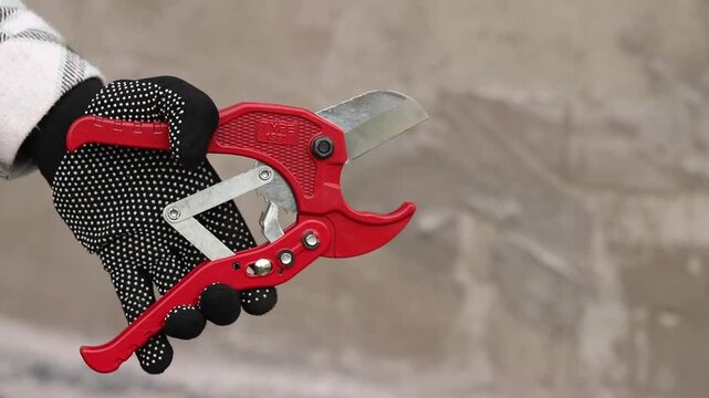 A hand in a work glove holds a red pipe cutter for plastic pipes. A hand holds professional scissors for cutting PVC and polypropylene pipes. A red tool with a sharp steel blade