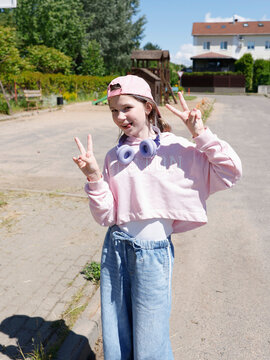 Trendy gen alpha girl in pink crop top hoodie showing peace sign. Cool tween wearing purple wireless headphones posing on sunny street. Youth street fashion lifestyle look during bright summer day