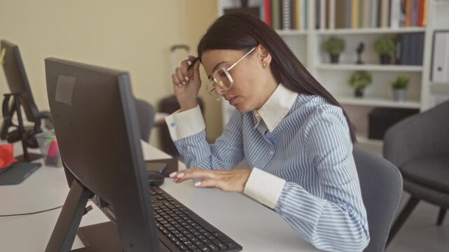 Young woman typing on keyboard while touching glasses at temple beside monitor and desk in office building; concentration focus.