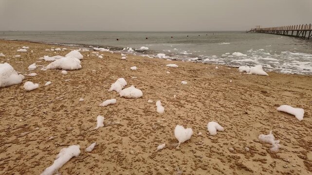 White foam patches on a quiet sandy beach with a long wooden pier and calm sea under a hazy sandstorm sky, summer travel in Egypt, Red Sea coast, UGC lifestyle