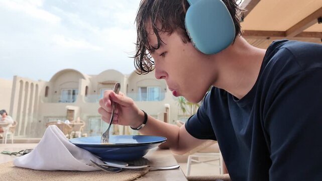 Fifteen year old teenage boy wearing large headphones eating pasta from a blue bowl at a sunny outdoor resort terrace, summer family vacation in Egypt, UGC travel lifestyle