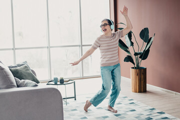 Happy senior woman dancing barefoot in bright living room with headphones, plants and cozy furniture enjoying music and a carefree moment at home