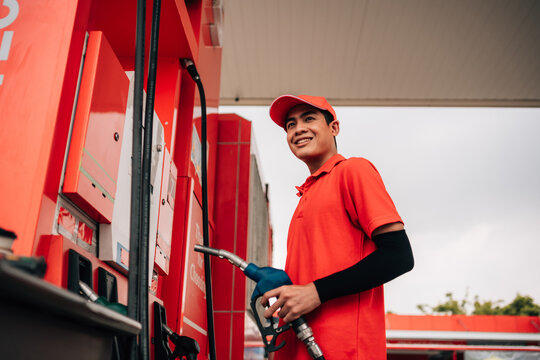 A bright, low-angle shot of a friendly young male gas station worker or service attendant dressed in a vibrant red polo shirt, matching red baseball cap, and black protective arm sleeves.  