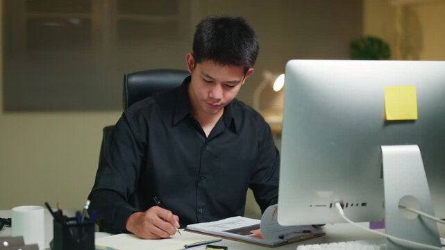Young Asian Businessman Working Late at Office Desk with Computer, Writing Notes in 4K