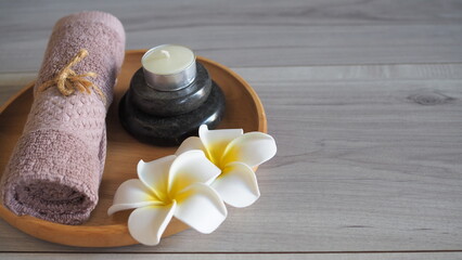 Minimalist zen spa still life with a soft mauve pink towel tied with rope, a scented candle on stacked black massage stones, and white frangipani flowers in a circular wooden tray. Set on a light gray © kanjana