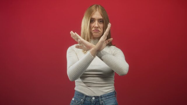 Woman with crossed hands forming an x, showing palms and a pouting expression in a red studio wearing white sweater and jeans; defiance.