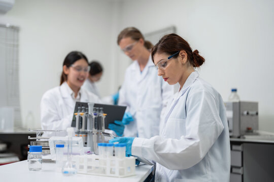 Professional female medical scientist reviewing clinical trial data on a digital tablet with diverse teamwork colleagues working in the background of a modern commercial pharmaceutical lab.