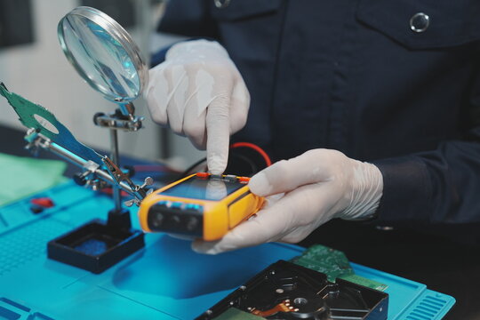Close-up of a technician interacting with a yellow digital multimeter next to a magnifying glass stand, adjusting the device to measure electrical currents on the hardware setup.