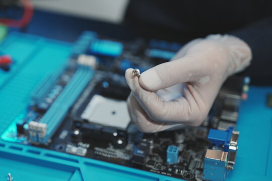 A close-up shot focused on a technician's gloved hand holding a small metal screw directly above a computer motherboard, preparing for hardware installation or assembly.