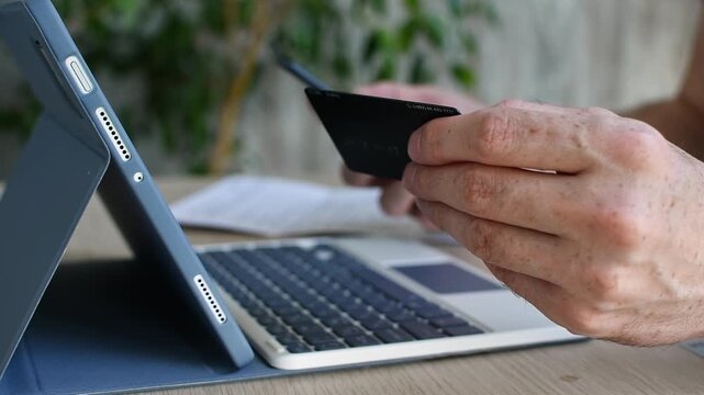 Person holds smartphone and credit card for online payment near tablet keyboard at home desk, close up of mobile banking