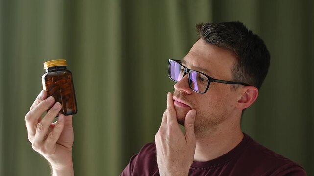 Thoughtful man examines unlabeled capsule bottle at home, considering health supplement, treatment choice, or daily routine