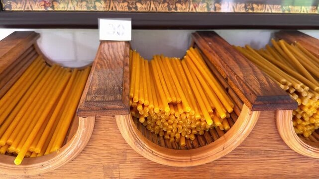 Assortment of wax candle with price tags in a wooden stand inside a church shop. Orthodox ritual supplies for believers and parish worship. Traditional beeswax candle in a temple counter.