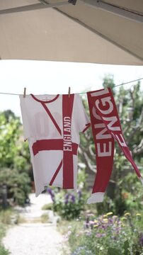 English football fan t-shirt and scarf hanging outdoors on clothesline under parasol drying in the wind, natural garden with flower beds blurred in background

