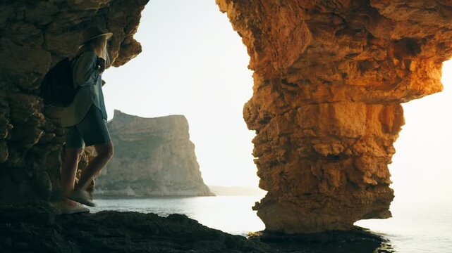 Young female traveler with backpack standing inside sea cave and admiring calm ocean during sunset, representing female freedom, independence, solo travel, and adventurous summer lifestyle.