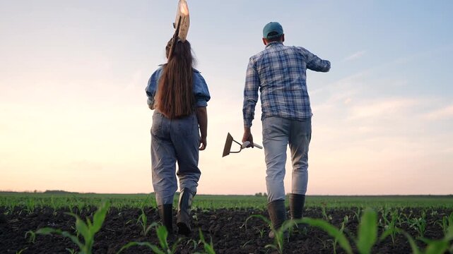A woman and a man walk through a green field at sunset. The man waves while holding a tool, and the woman carries a hoe as they leave the farm.