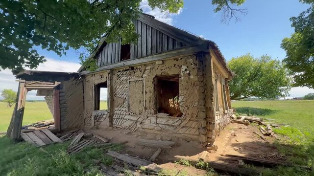 The dilapidated house stands alone in a wide field, surrounded by trees and open grass. The old house shows signs of decay, with broken walls and a weathered roof.
