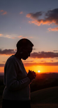 Image of a woman in silhouette praying at sunset with her hands clasped together