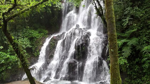 Aerial drone view of a wide multi-tiered waterfall cascading over dark volcanic rock surrounded by lush tropical jungle foliage, Hidden Falls, Bulusan, Sorsogon, Philippines.