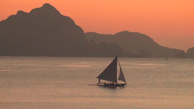 Outrigger sailboat moving through quiet island water with mountains behind