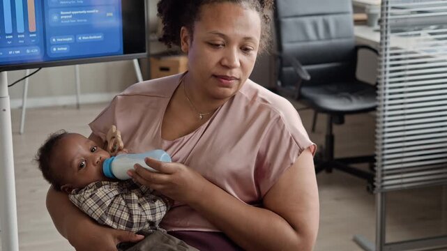 African American mother holding baby and feeding with bottle while working in office. Ideal for commercial videos highlighting work-life balance, remote work, and modern parenting