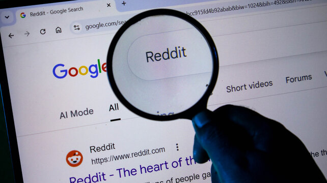 Closeup of a hand holding a magnifying glass over a computer screen showing a google search result for reddit
