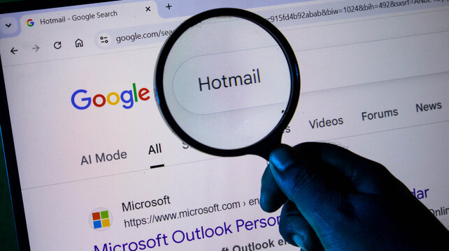 Closeup of a hand holding a magnifying glass over a computer screen showing a google search for hotmail