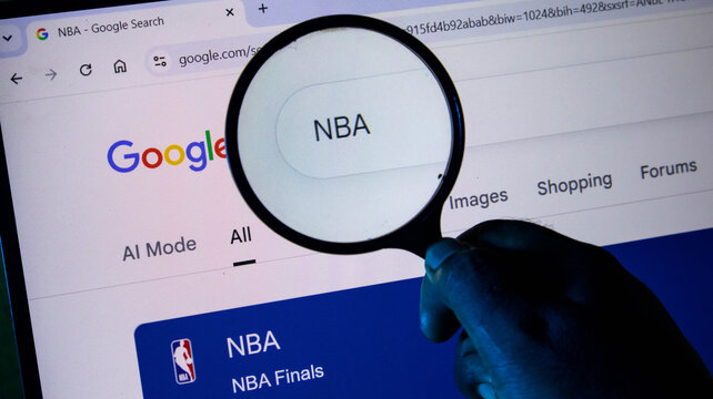 A hand holding a magnifying glass over a computer screen showing a google search for nba, emphasizing digital research and sports information