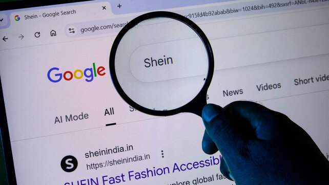 A hand holding a magnifying glass over a computer screen displaying a google search result for the online fashion retailer shein