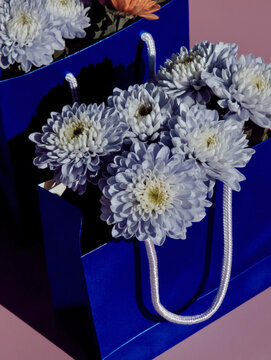 White chrysanthemums overflowing from a blue gift bag on a pink studio background. Floral gift and greeting card concept for stationery, seasonal branding, social media, and packaging.