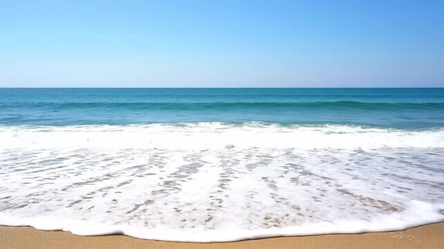 A serene beach scene showing foamy waves rolling onto the sand with the vast blue ocean in the background under a clear, cloudless sky