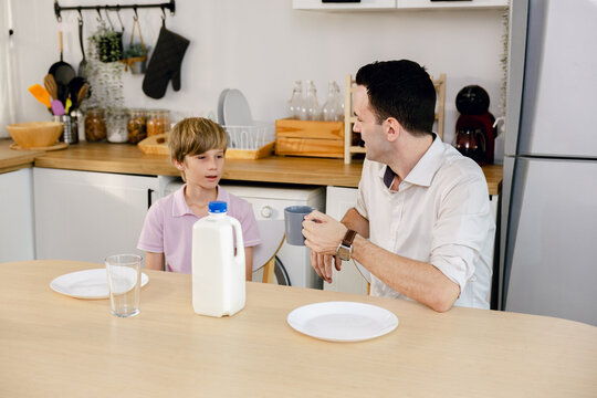 A father and son sit together at a kitchen table with milk and empty plates, sharing a quiet moment in a modern home.